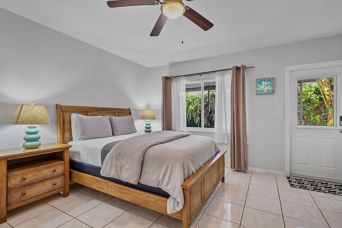 Une chambre à coucher avec un lit en bois, deux tables de chevet, un ventilateur de plafond, une fenêtre avec des rideaux et une porte avec un paillasson.