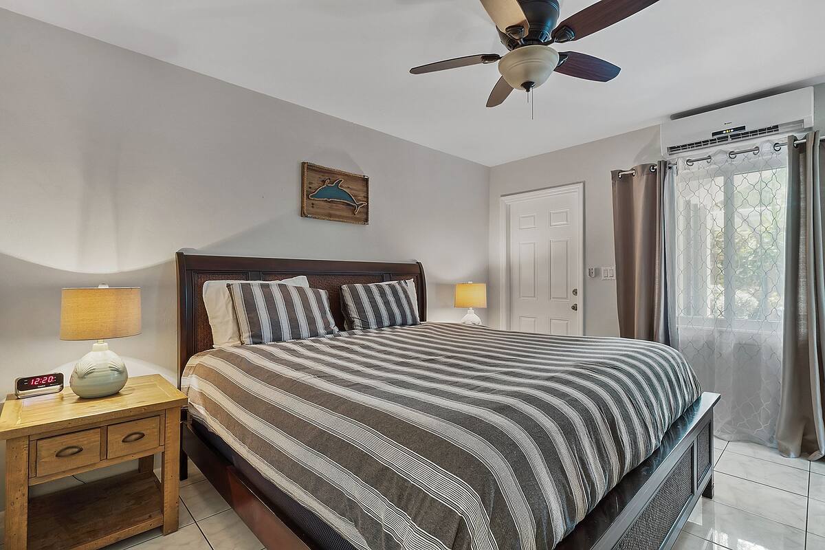Une chambre à coucher comprenant un lit, des tables de chevet, un ventilateur de plafond, une horloge murale et une fenêtre avec des rideaux.