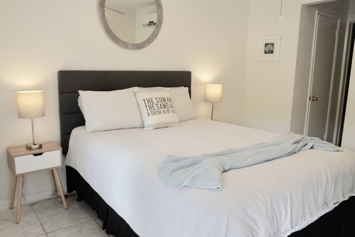 Un lit bien fait, avec une couette blanche et deux oreillers, dont l’un porte un motif. On aperçoit une table de chevet avec une lampe, un miroir fixé au mur et une porte sur la droite.