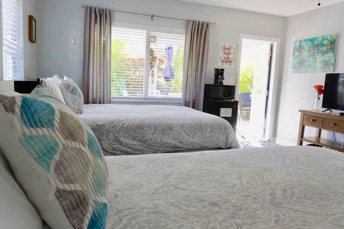 Une chambre avec deux lits, un téléviseur posé sur un meuble en bois et une fenêtre équipée de stores.