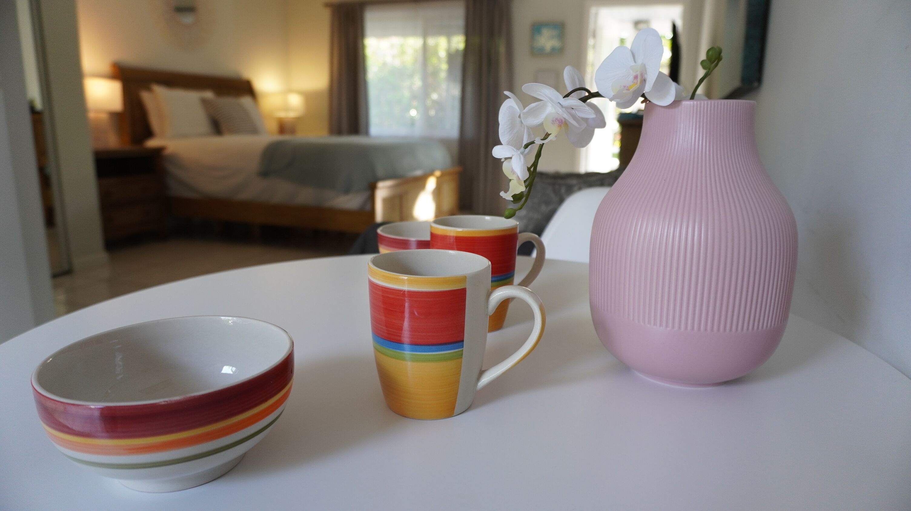 Habitación de hotel con cama, mesita de noche, lámpara y ventana con cortinas. Se observa una mesa con un florero rosa, un tazón y dos tazas.