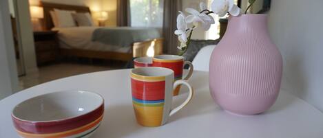 Habitación de hotel con cama, mesita de noche, lámpara y ventana con cortinas. Se observa una mesa con un florero rosa, un tazón y dos tazas.