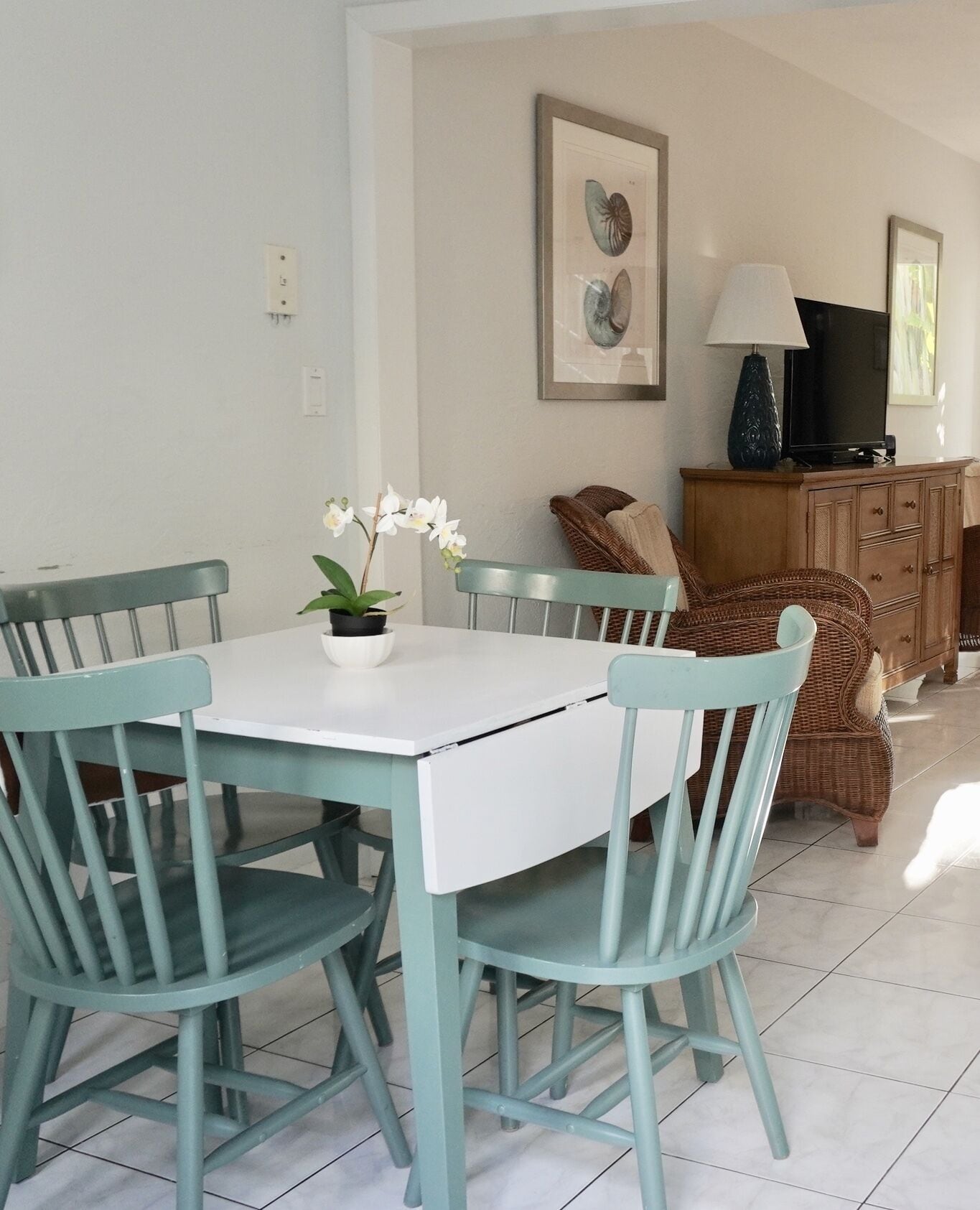 Un comedor con una mesa blanca y sillas color turquesa, una orquídea en maceta y muebles de mimbre.