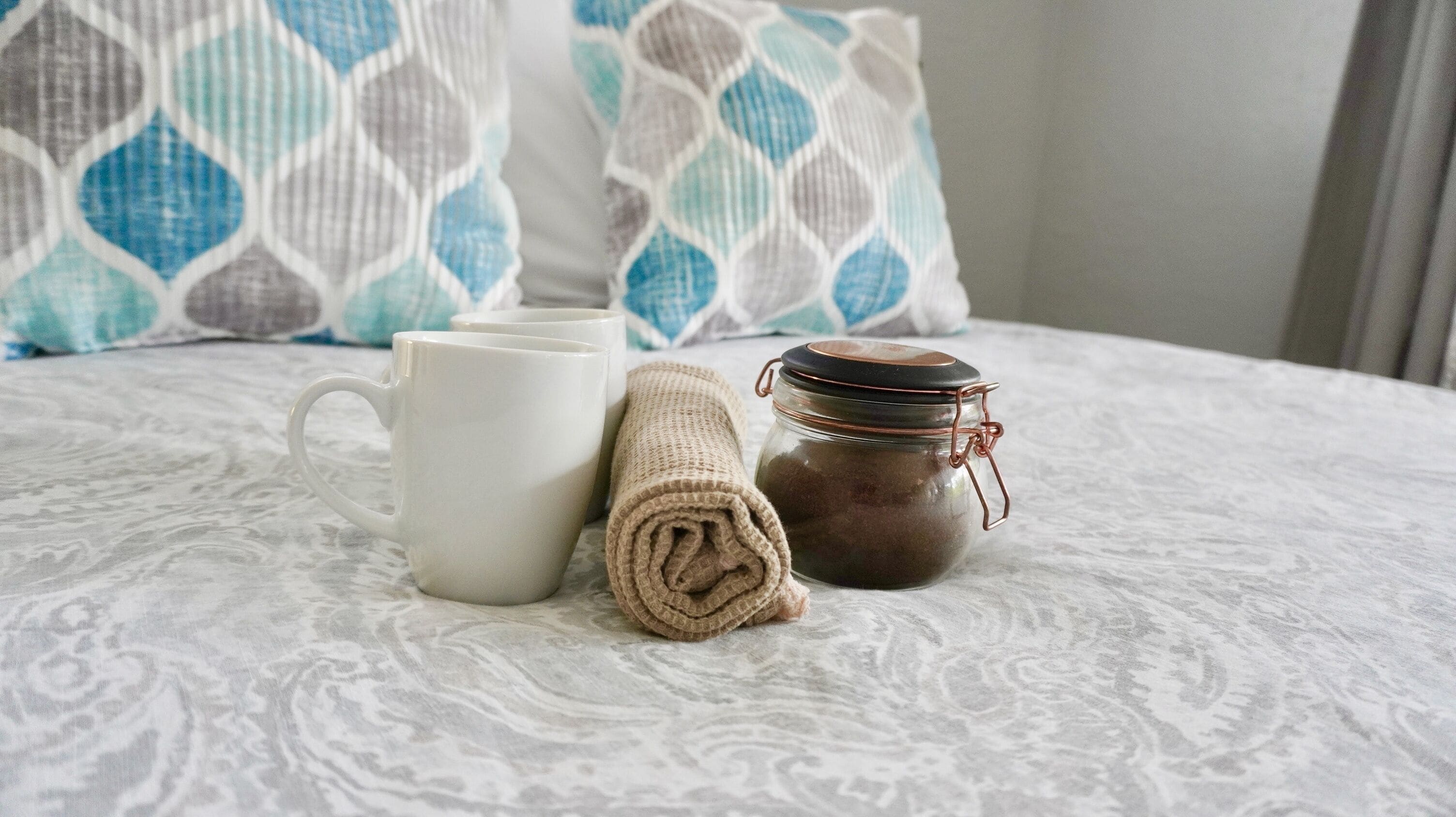 Un lit avec une couvre-lit à motifs, une serviette enroulée, une tasse blanche et un bocal avec un couvercle en métal.