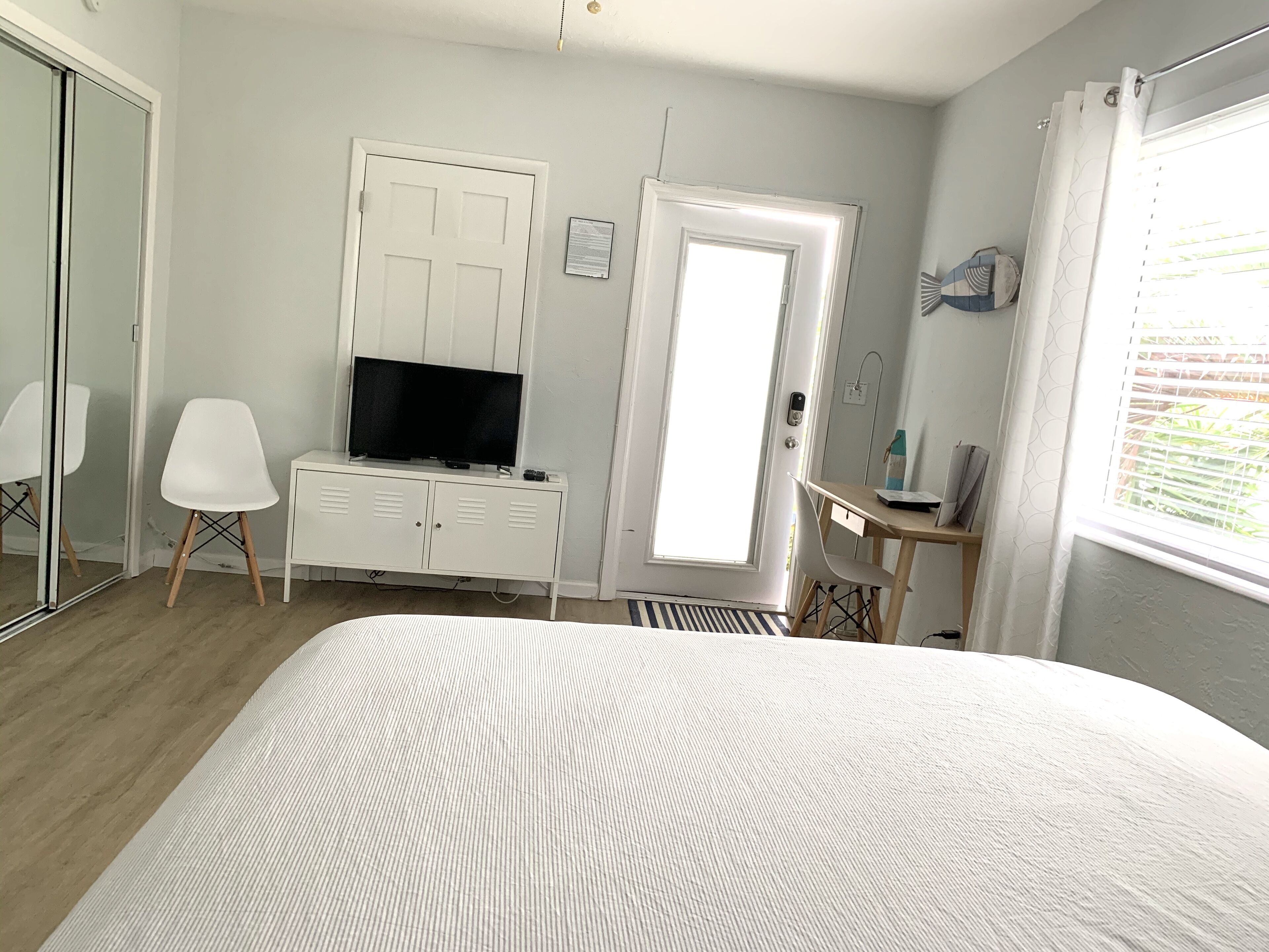 A hotel room with a bed, a TV on a white dresser, a desk with a chair, a door, and a window with blinds.