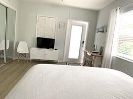 A hotel room with a bed, a TV on a white dresser, a desk with a chair, a door, and a window with blinds.