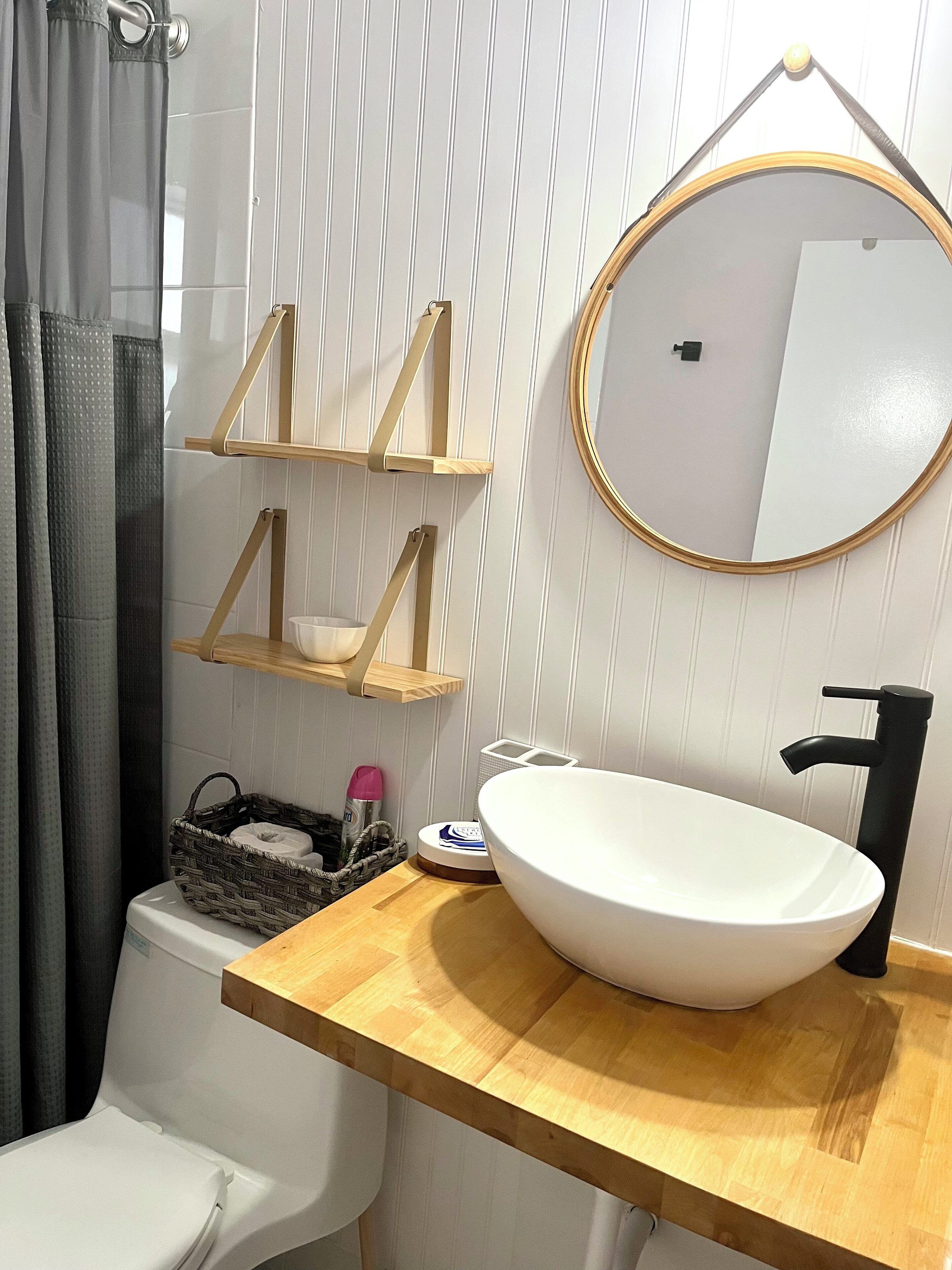 A bathroom with a white sink, black faucet, and a round mirror on the wall. There are wooden shelves above the sink and a basket on the floor.
