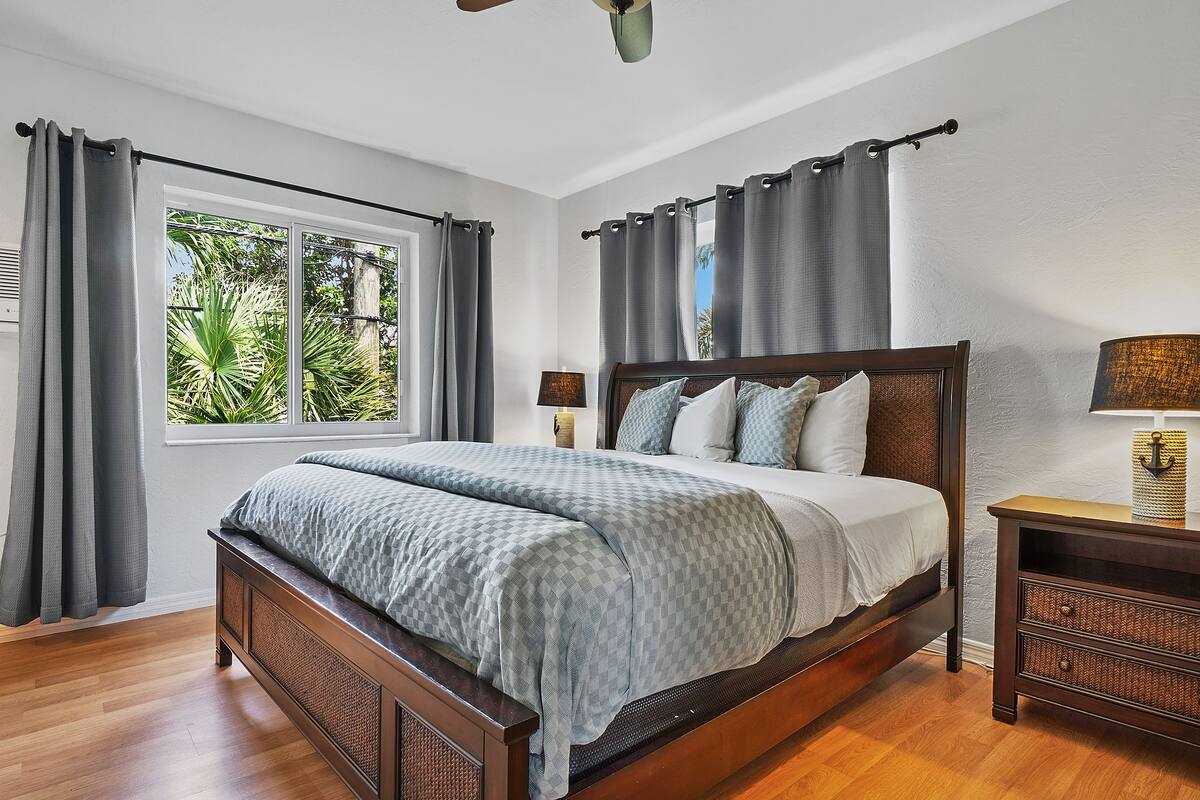 Une chambre à coucher avec un grand lit, une table de chevet, une lampe et une fenêtre avec des rideaux.