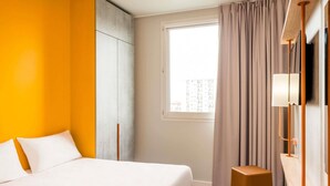 Habitación de hotel con cama, un taburete pequeño, ventana con cortinas y televisor montado en la pared.