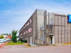 A modern building with a metal facade, a blue sign, and a parking lot. - ibis budget Metz Technopole (Metz)