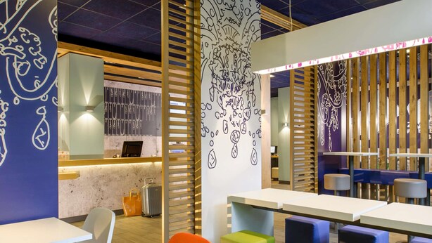 Un interior moderno con mesas blancas y taburetes coloridos, una partición de listones de madera y una pared con diseños abstractos.