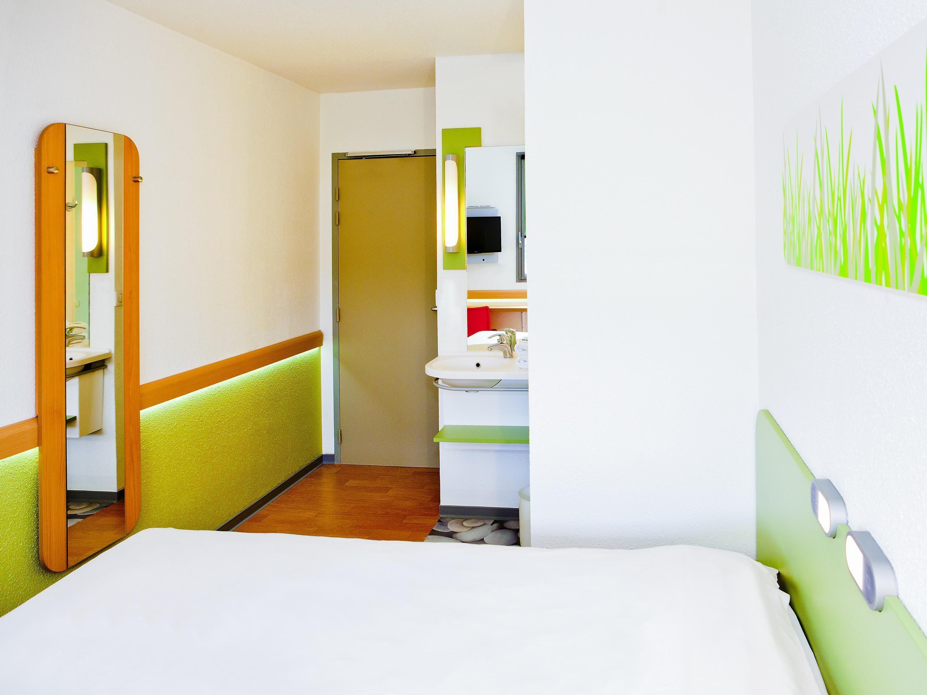 Una camera d'albergo con un letto, un bagno con lavandino e specchio, e un corridoio con una porta.