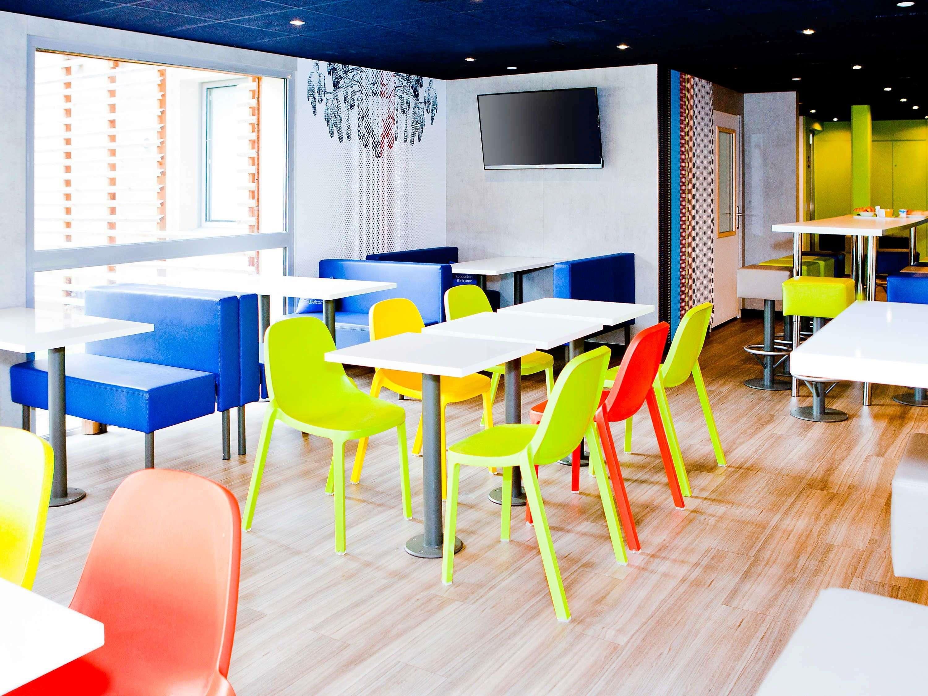 Ein modernes Restaurant-Interieur mit bunten Stühlen und Tischen, einem Flachbildfernseher an der Wand und einem großen Fenster mit Jalousien.