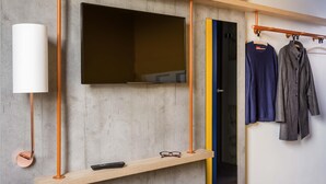 Una habitación moderna con un televisor montado, un estante de madera con un control remoto y un perchero con dos prendas.