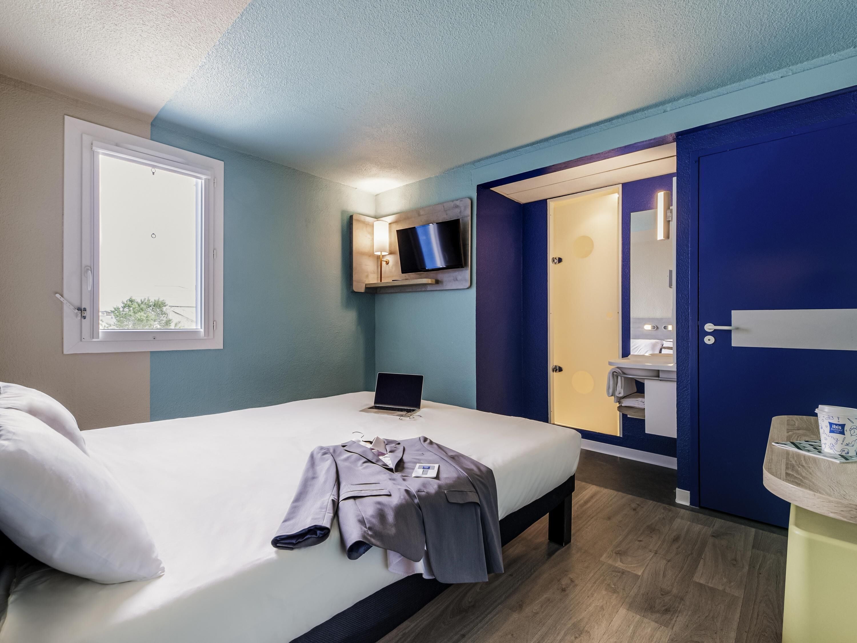 Habitació d'hotel amb un llit, un ordinador portàtil, un abric i un bany amb mirall i un lavabo.
