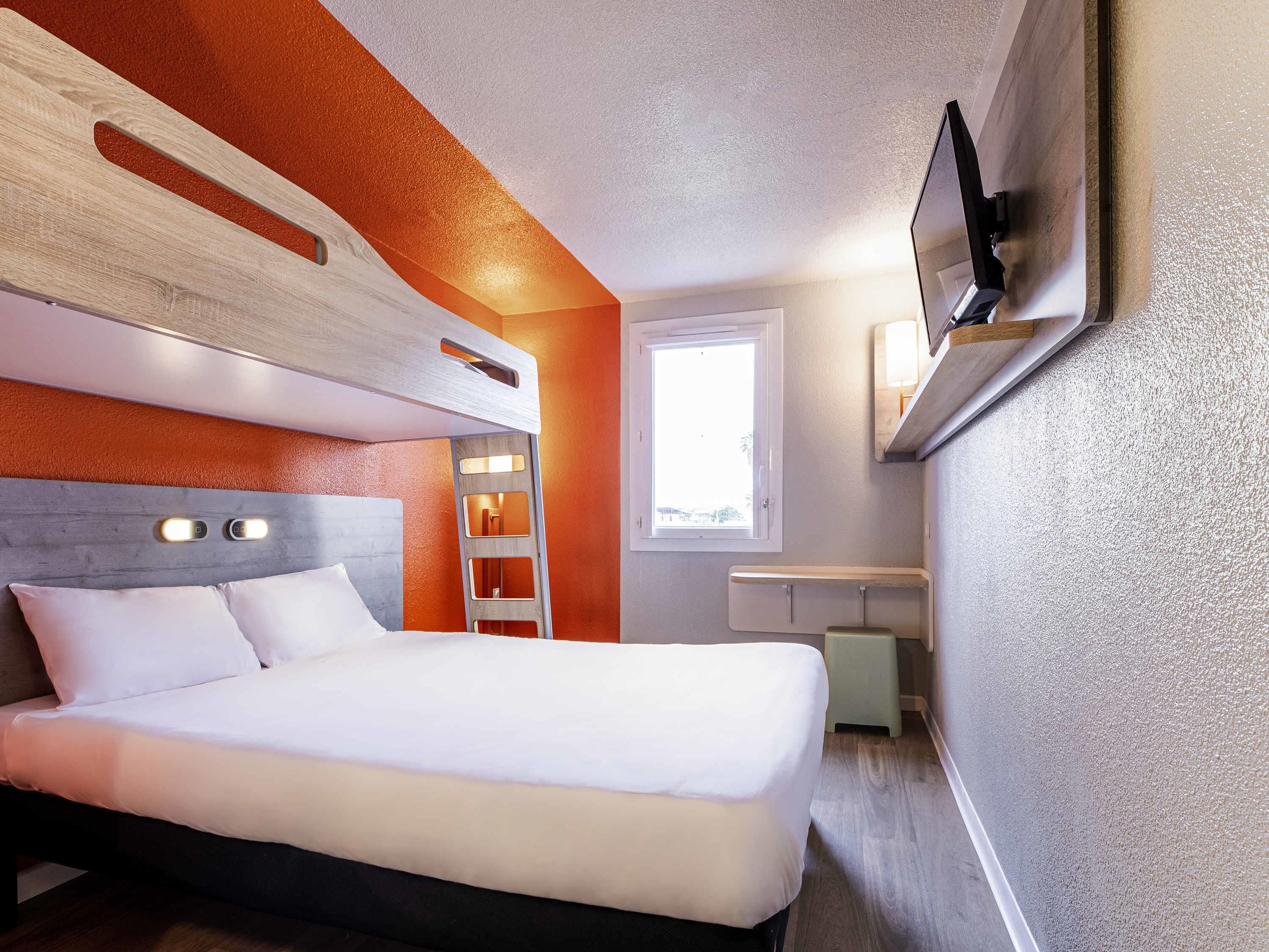 Ein modernes Hotelzimmer mit einem Bett, einem an der Wand befestigten Fernseher, einem Fenster und einer fest eingebauten Ablagefläche.