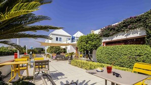 An outdoor seating area with yellow and grey chairs and tables, surrounded by greenery and a white building with a blue roof. - ibis budget Perpignan Sud (Perpignan)