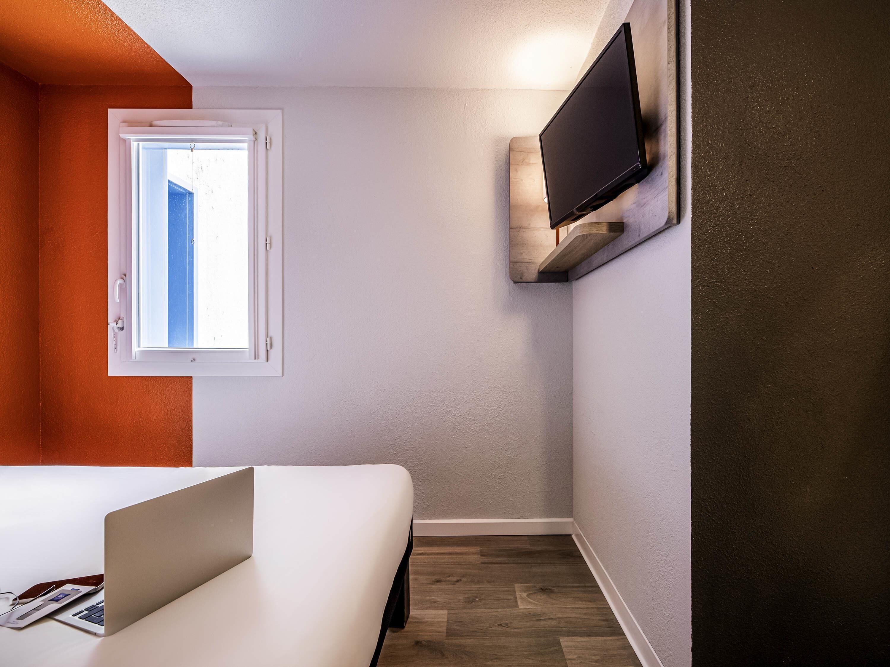 A room with a bed, a window, a mounted TV, and a laptop on the desk.