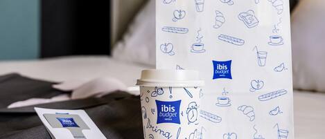 A hotel room with a bed, a white bag with blue doodles, and a coffee cup.