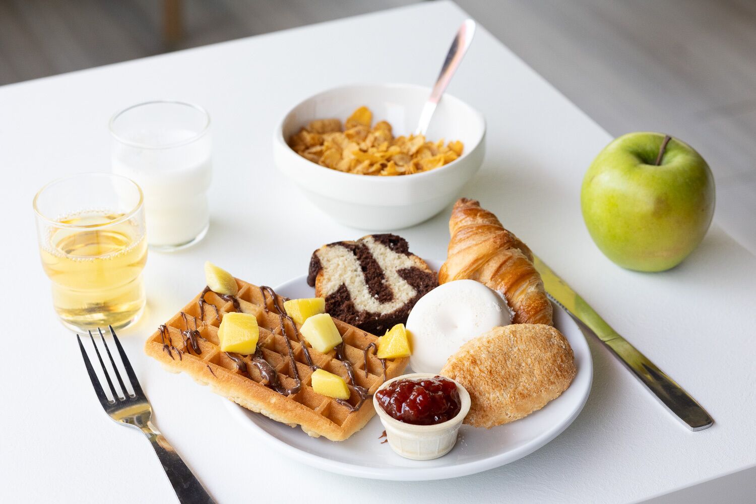 Une assiette de petit-déjeuner avec des gaufres, un croissant, une part de gâteau, une pomme verte, un verre de lait, un bol de céréales et une fourchette, le tout posé sur une table blanche.