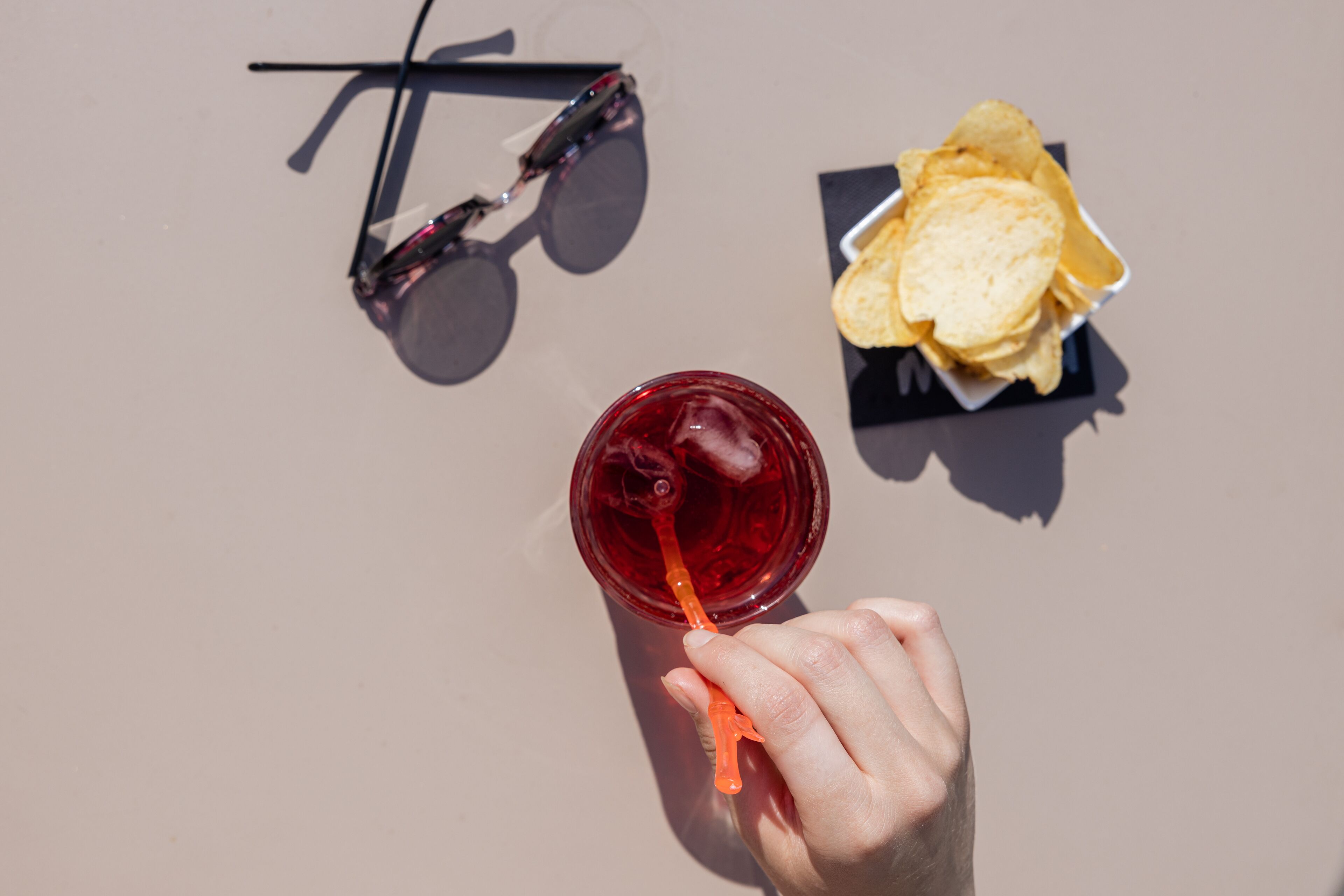 Una mano sostiene una bebida roja con pajita, gafas de sol y un cuenco pequeño de patatas fritas sobre una superficie plana.