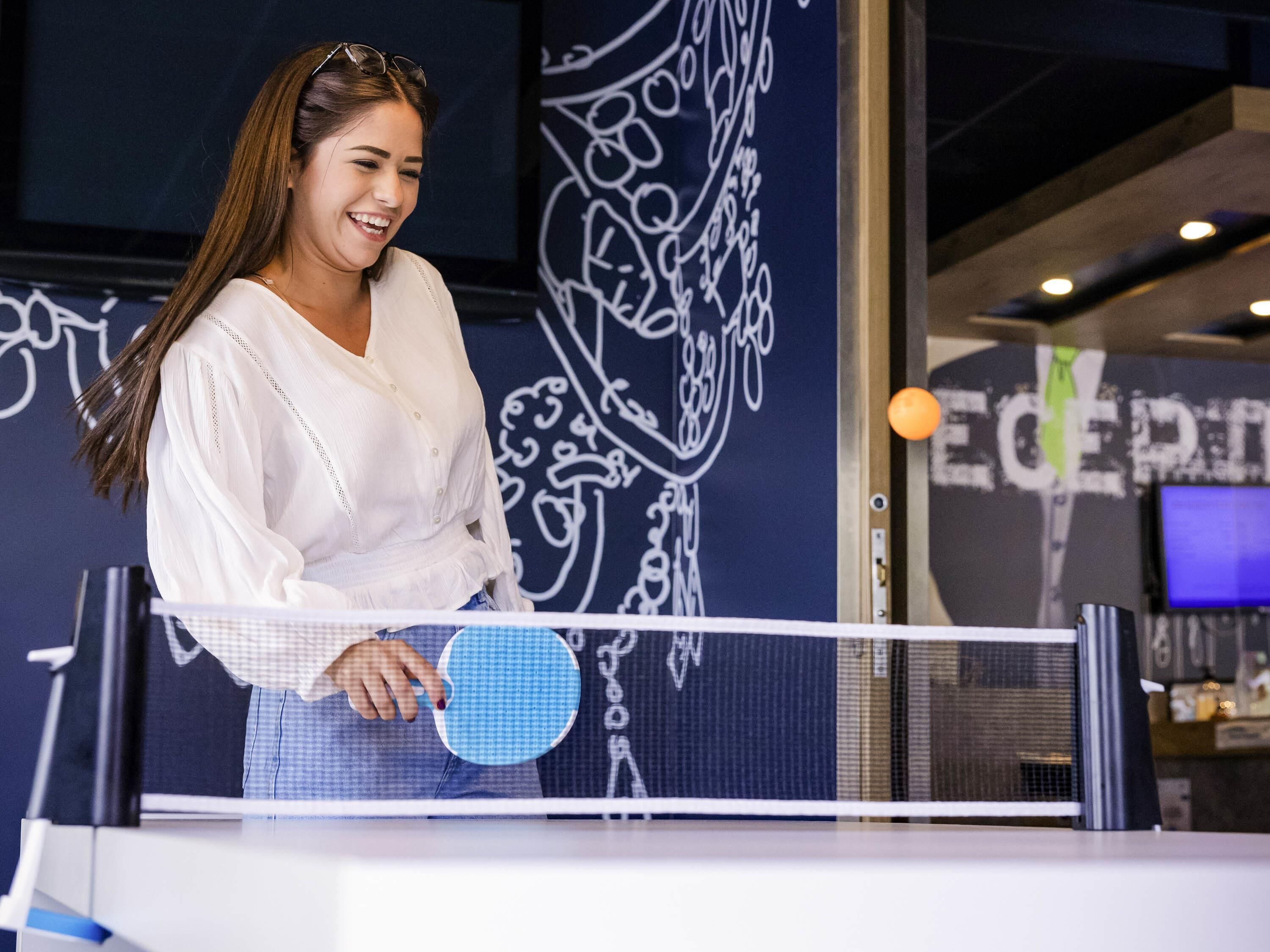 A person holding a paddle, standing in front of a table tennis table with a wall featuring abstract designs in the background.