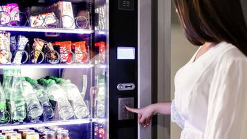 Máquina expendedora llena de aperitivos y bebidas, una mujer interactuando con ella, y una pantalla digital.
