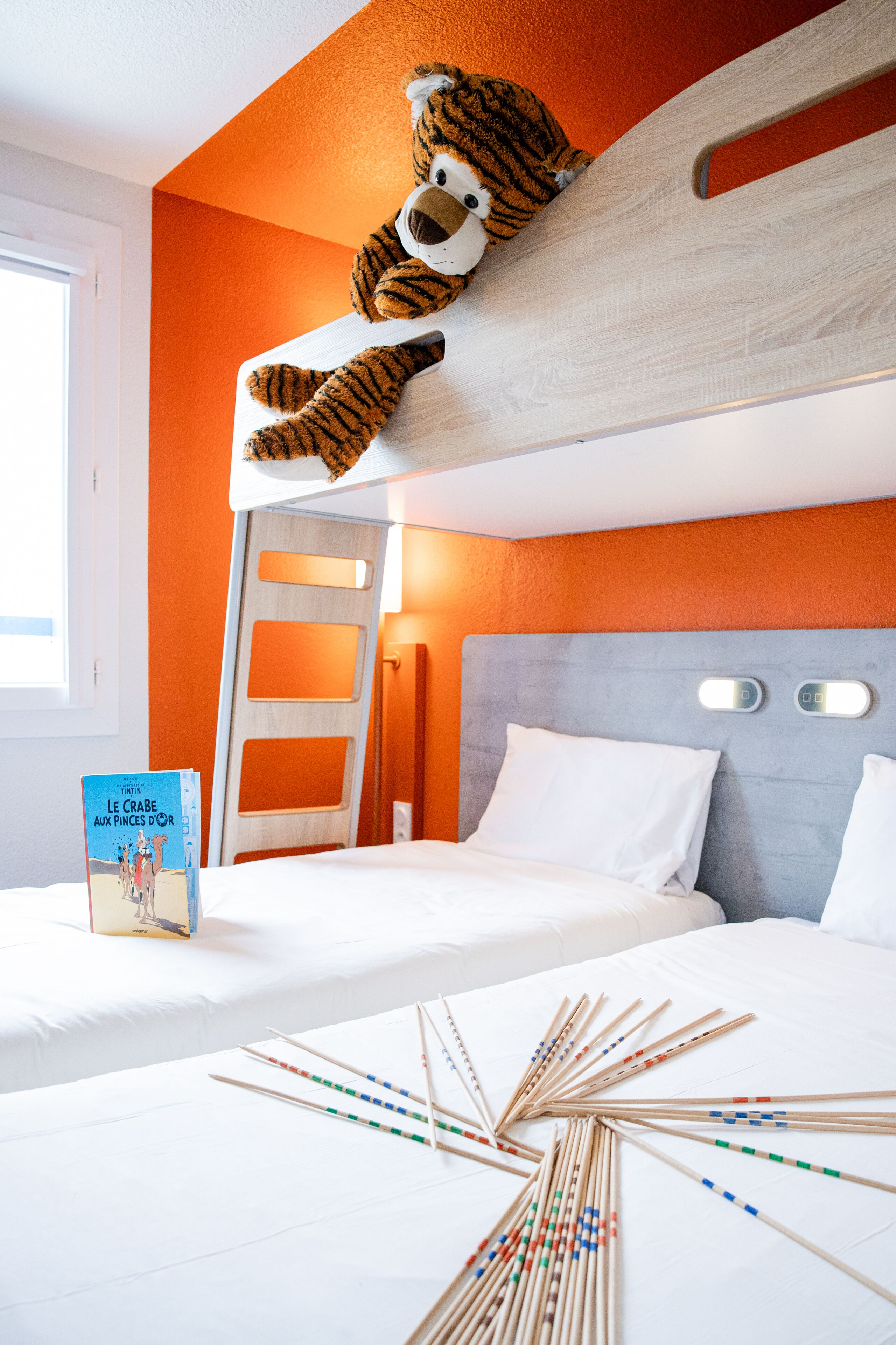 Una habitación de hotel con una litera, un oso de peluche en la parte superior, un libro sobre la cama y un juego de palillos chinos dispuestos sobre la cama.