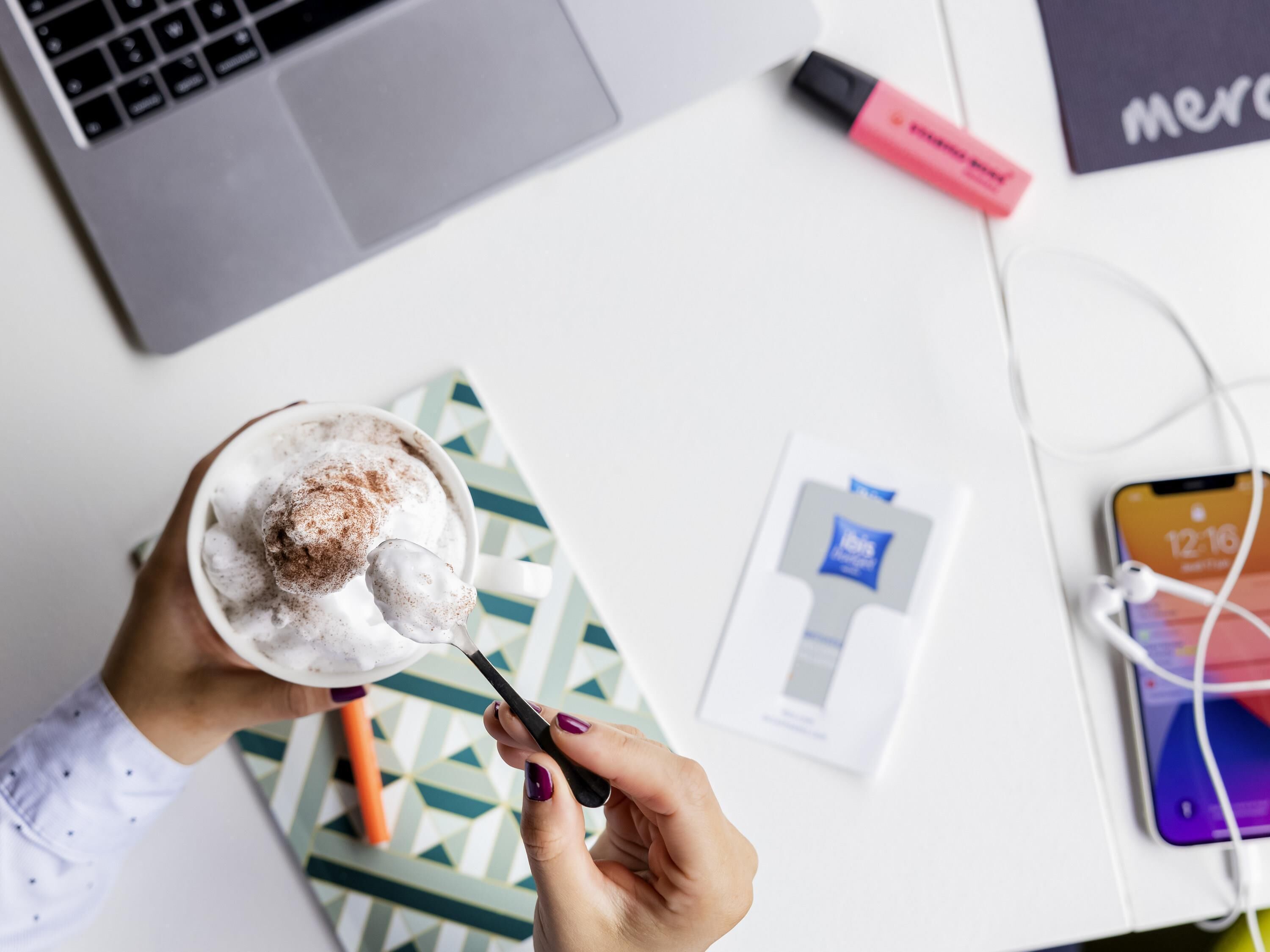 A person holding a cup with a spoon, a laptop, a smartphone, and a pen on a desk.