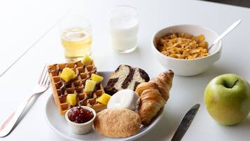 A breakfast plate with waffles, a croissant, a muffin, a bowl of cereal, a glass of milk, a glass of juice, a small bowl of jam, and a green apple.
