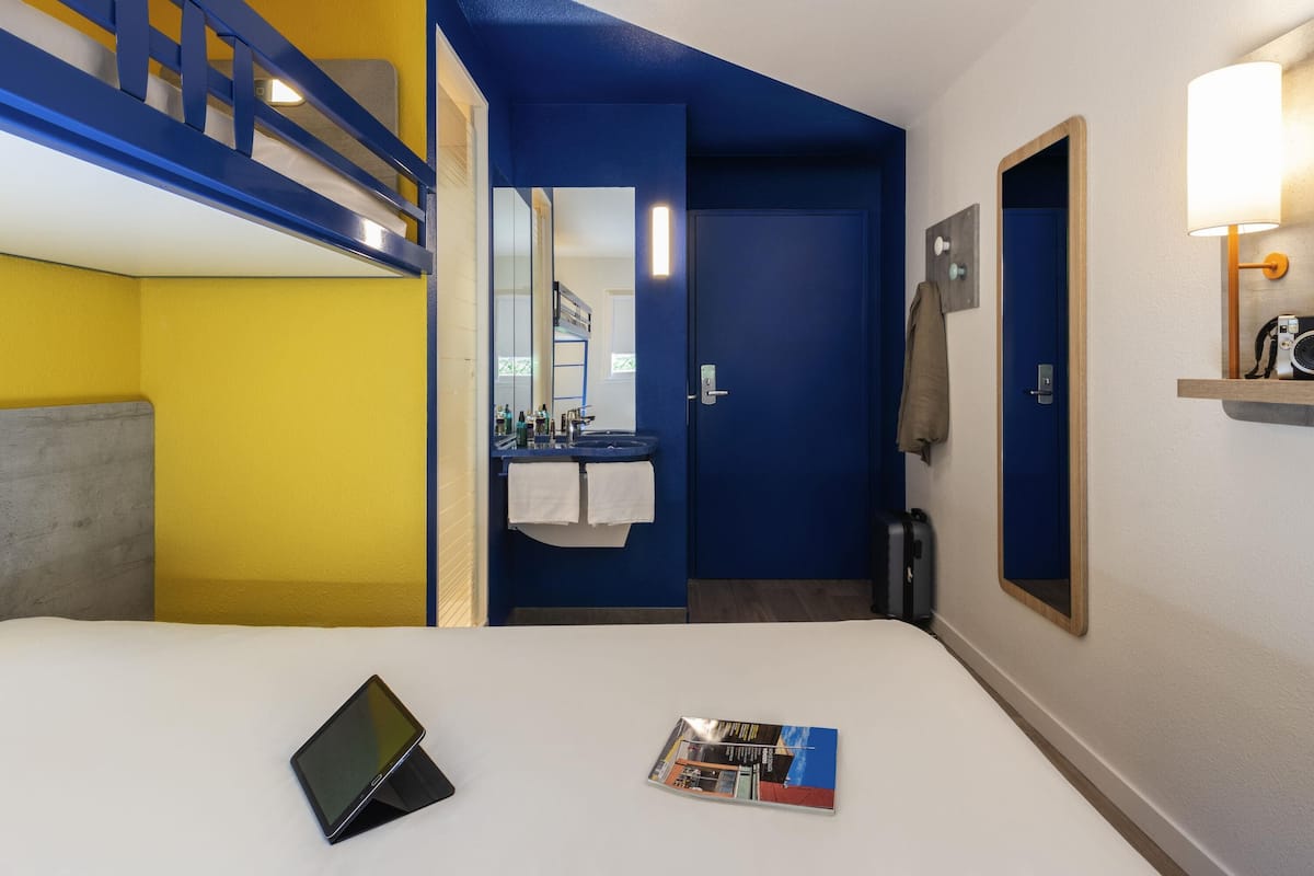 Une chambre d’hôtel avec un lit, une tablette, un magazine, une salle de bain avec un miroir et une porte bleue.