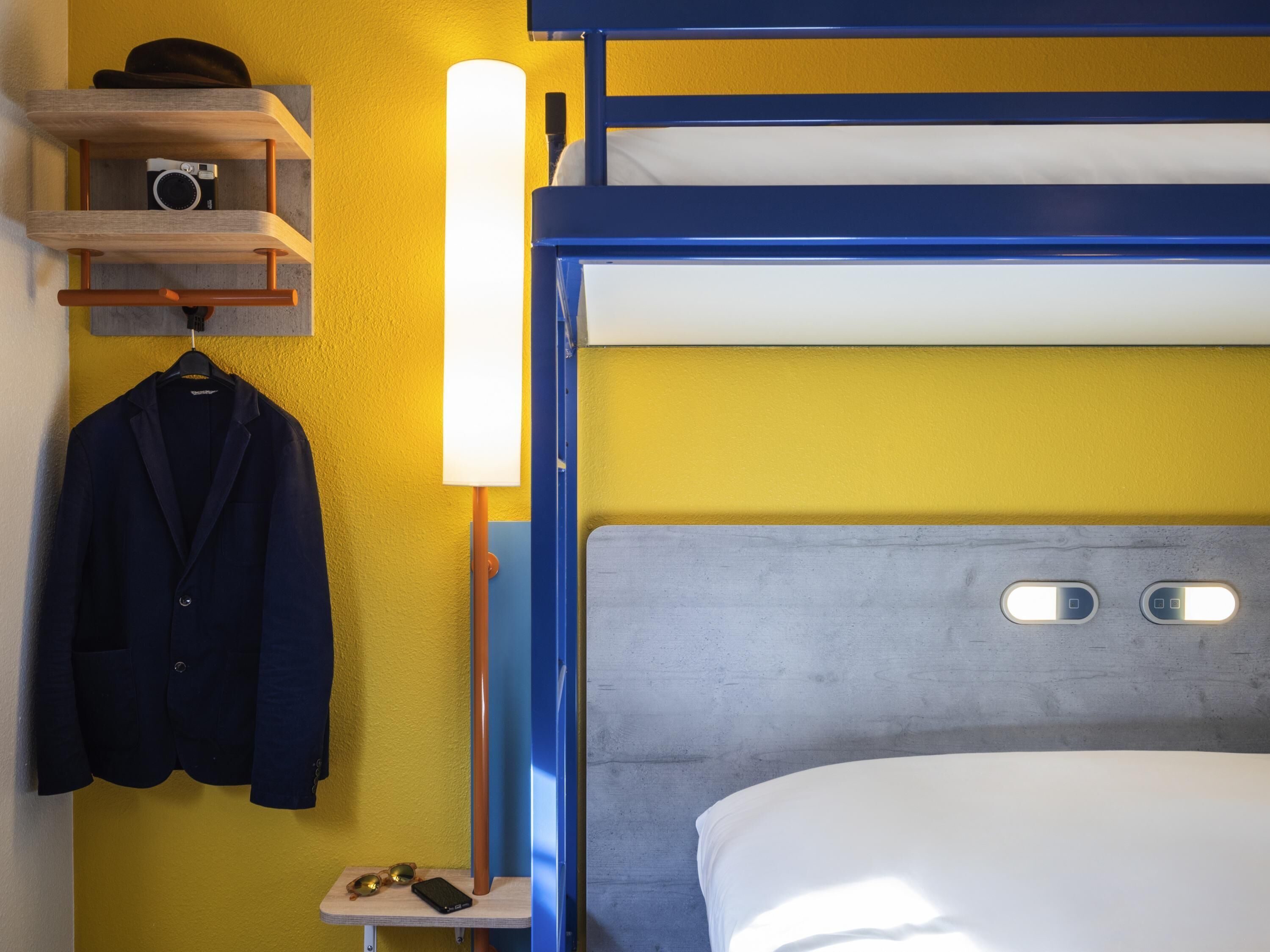 A bunk bed room with a wooden shelf holding a camera and a jacket, a bedside table with a lamp, and a bed with a grey headboard.