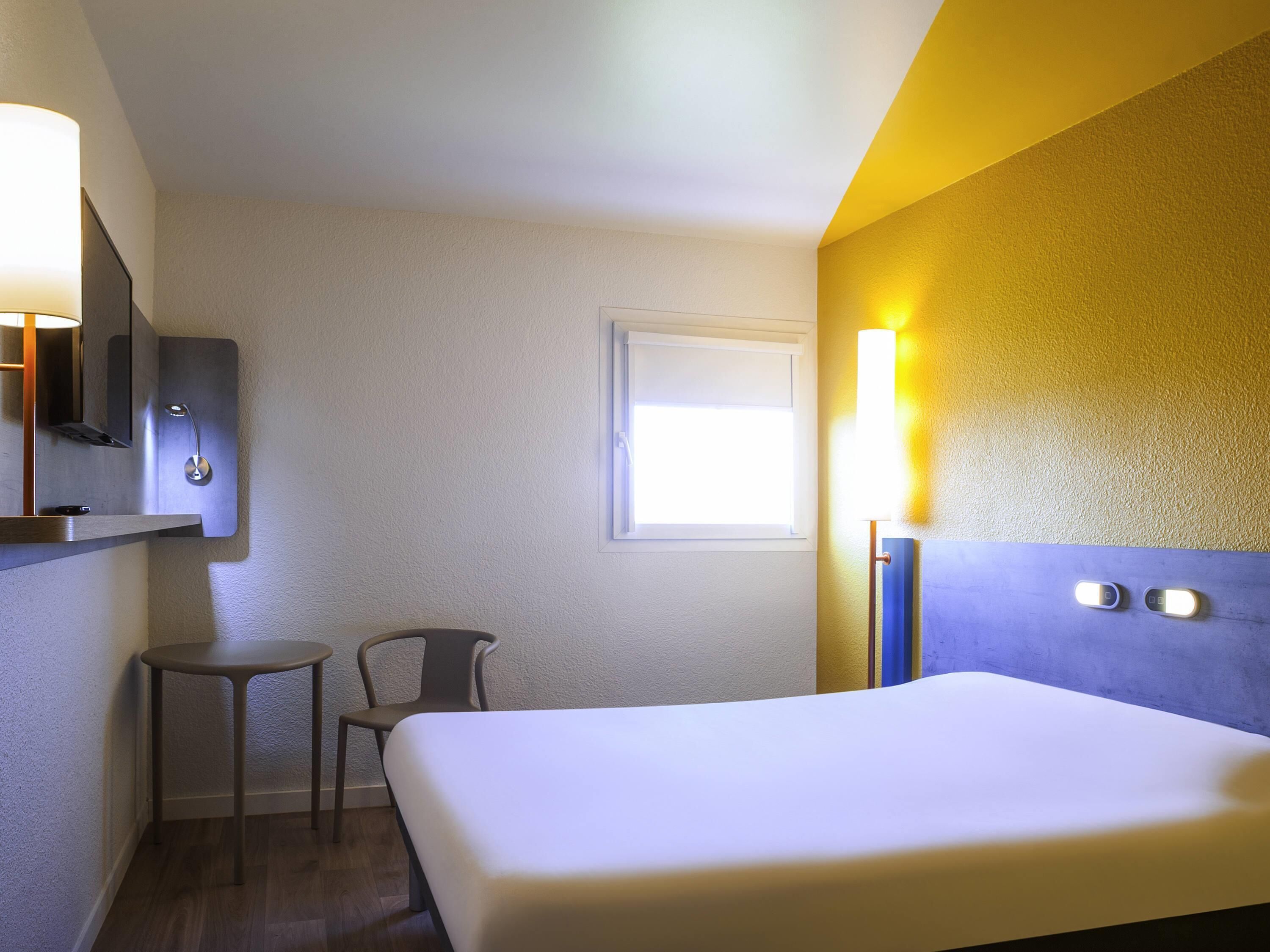 Une chambre d’hôtel comprenant un lit, une chaise, une petite table, un miroir, un téléviseur fixé au mur et une fenêtre.
