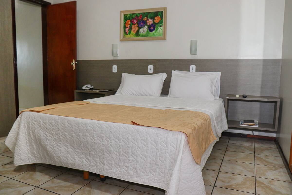 A neatly made bed with a white quilt and a beige throw blanket, a bedside table with a phone and a lamp, and a painting of flowers on the wall.