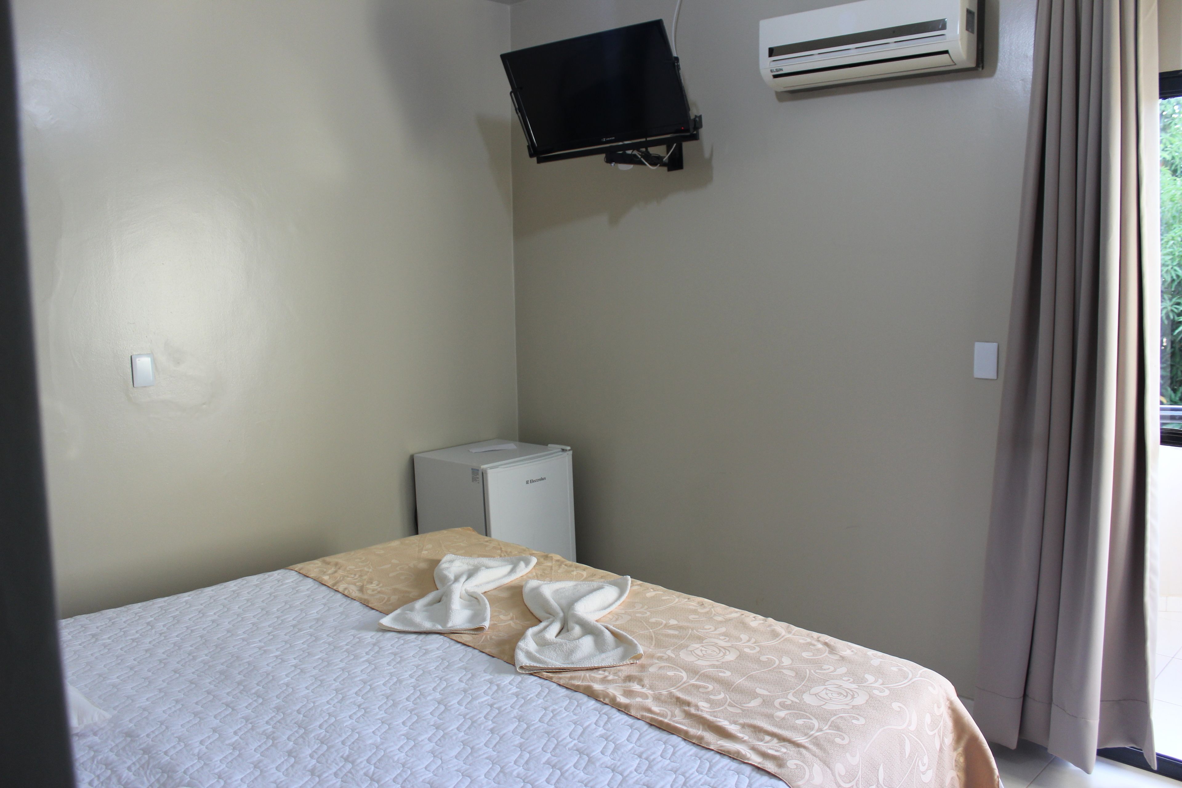 Una habitación pequeña y limpia con una cama, un televisor de pantalla plana montado en la pared y un equipo de aire acondicionado.