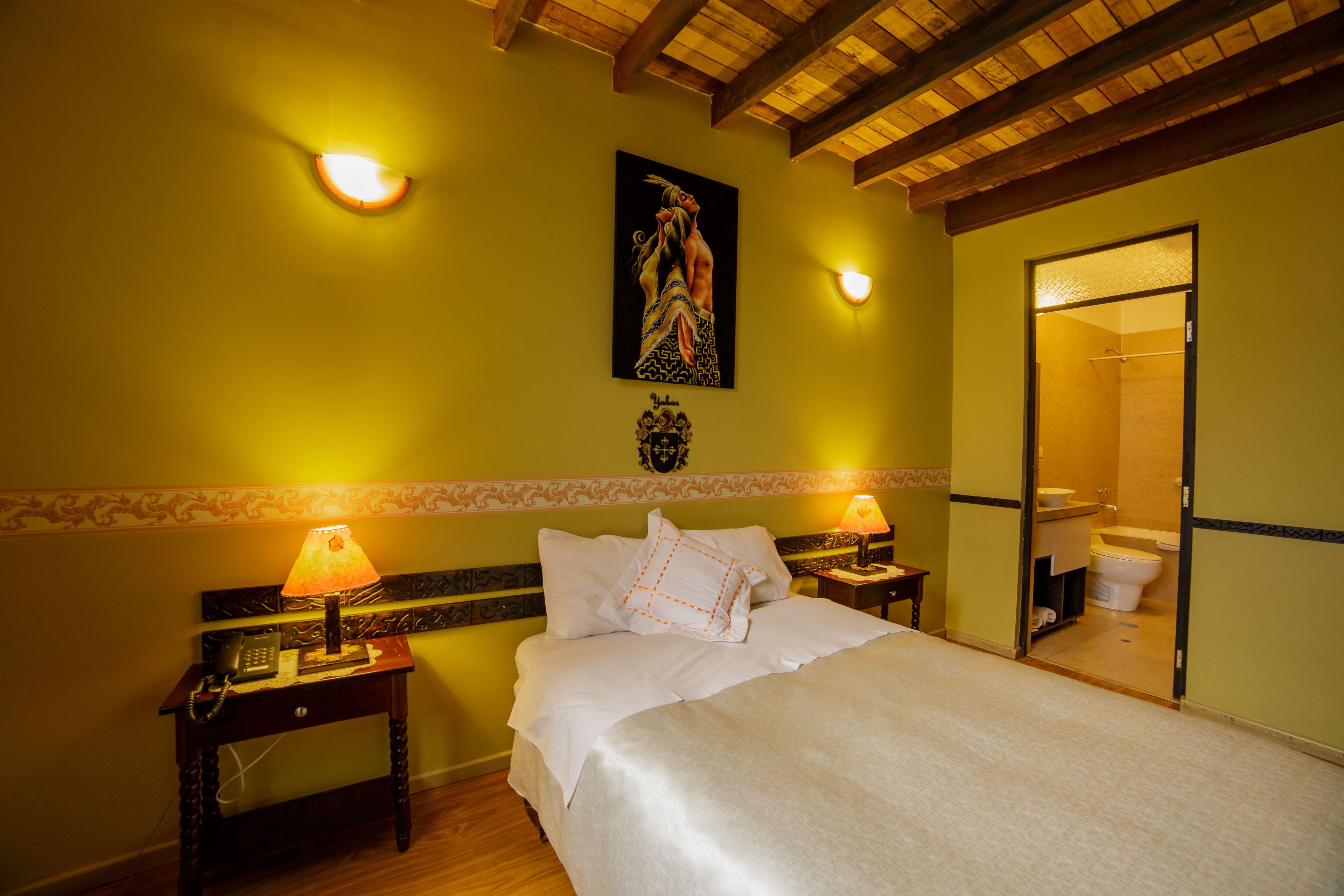 A hotel room with a bed, two bedside lamps, a painting on the wall, and a bathroom visible through an open door.