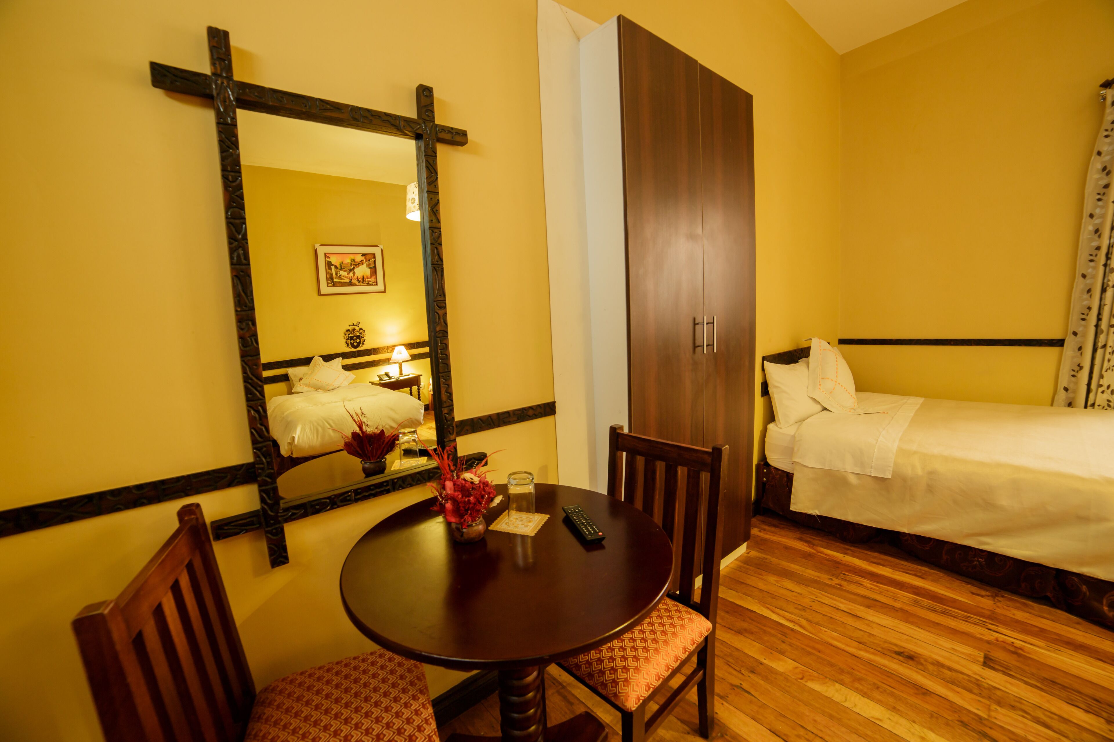 Habitación de hotel con piso de madera, una mesa redonda con sillas, cama con ropa blanca, armario y un espejo grande en la pared.