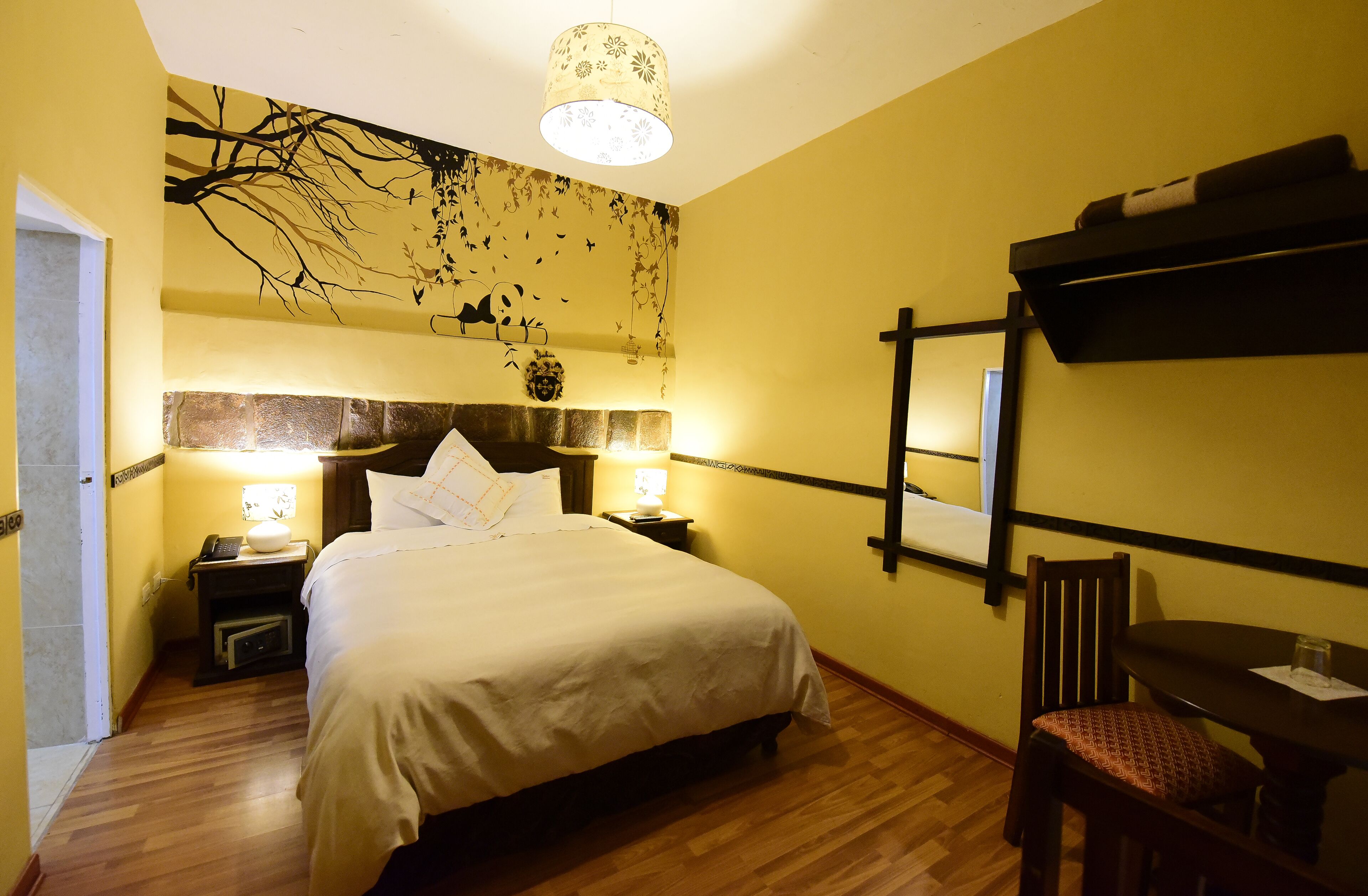 Habitación de hotel con cama, lámparas de noche, suelo de madera, estante montado en la pared y una mesa con una silla.