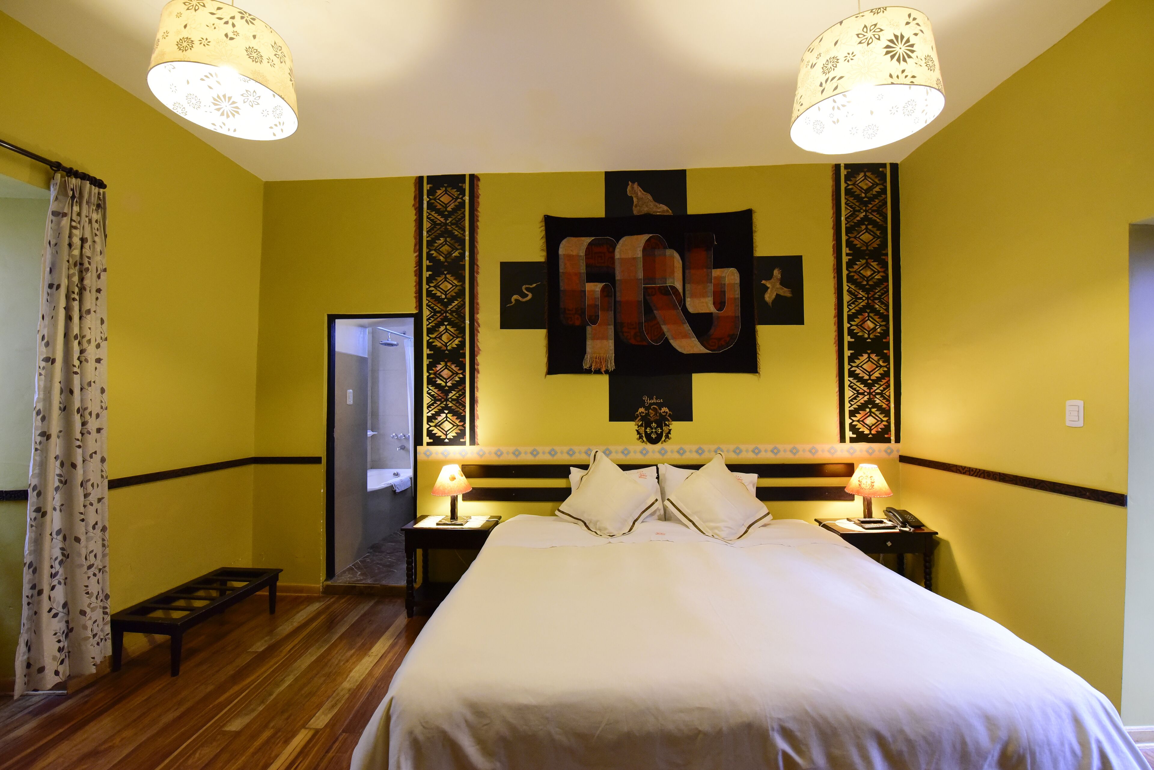 A hotel room with a large bed, two bedside lamps, a decorative wall hanging, and a wooden bench.