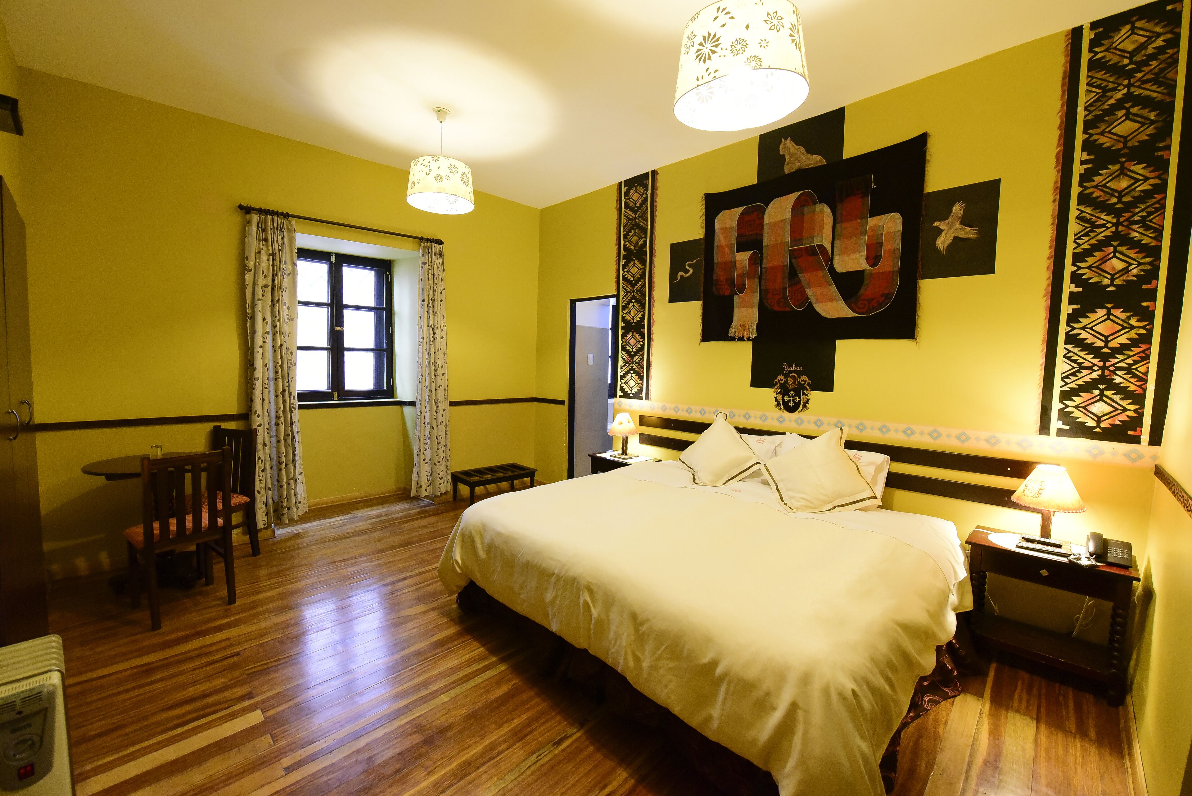 A hotel room with a large bed, a bedside table with a lamp, a window with curtains, and a wall with a geometric design.