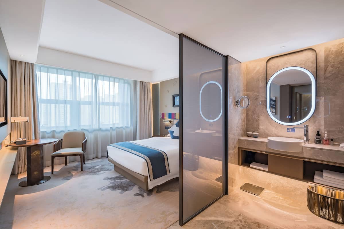 A modern hotel room with a large bed, a desk, a chair, a bathroom with a vanity and mirror, and a large window with curtains.