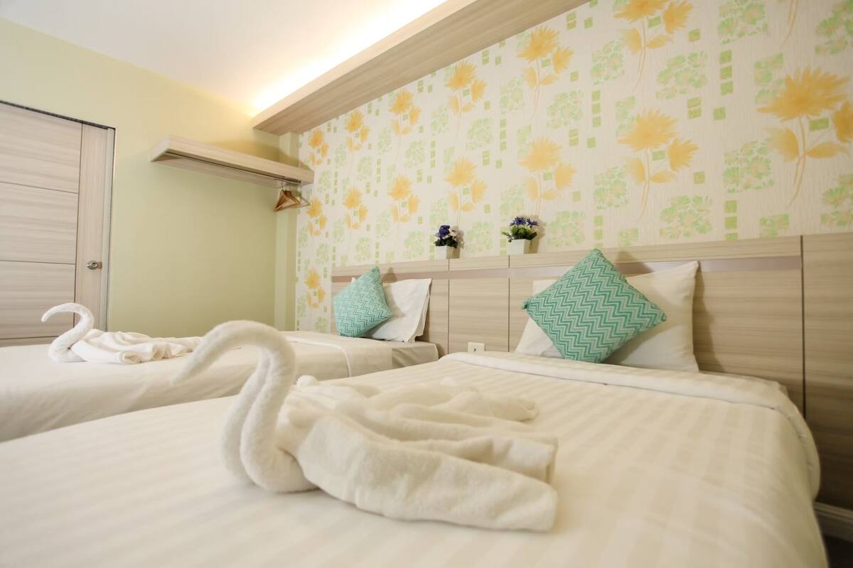 Une chambre d’hôtel avec deux lits, chacun recouvert d’une courtepointe blanche et agrémenté d’oreillers assortis. Des serviettes décoratives en forme de cygne sont posées sur les lits. La chambre a un mur de couleur vert clair orné d’un motif floral et une tête de lit en bois.
