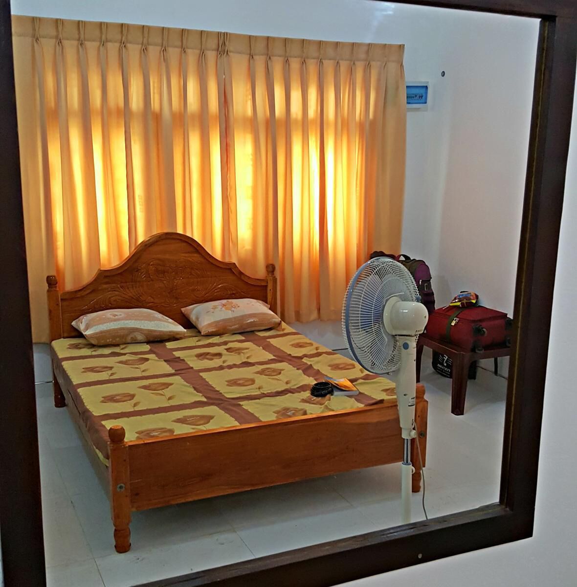 Un lit simple avec une tête de lit en bois, deux oreillers, une table de chevet avec un ventilateur et une valise posée sur le sol.