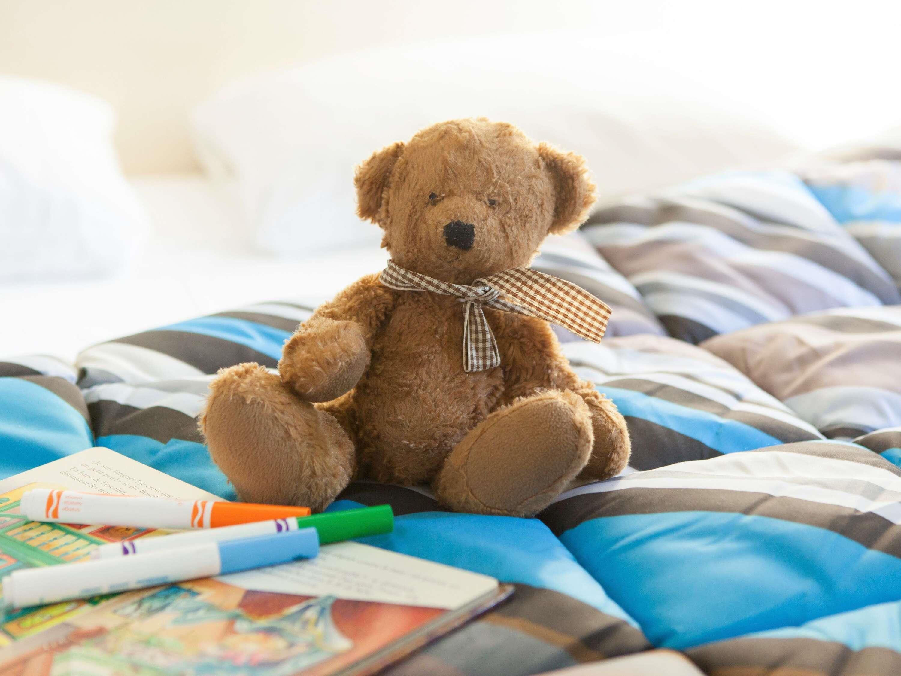 Un osito de peluche sobre una cama con ropa de cama a rayas y algunos libros y marcadores.