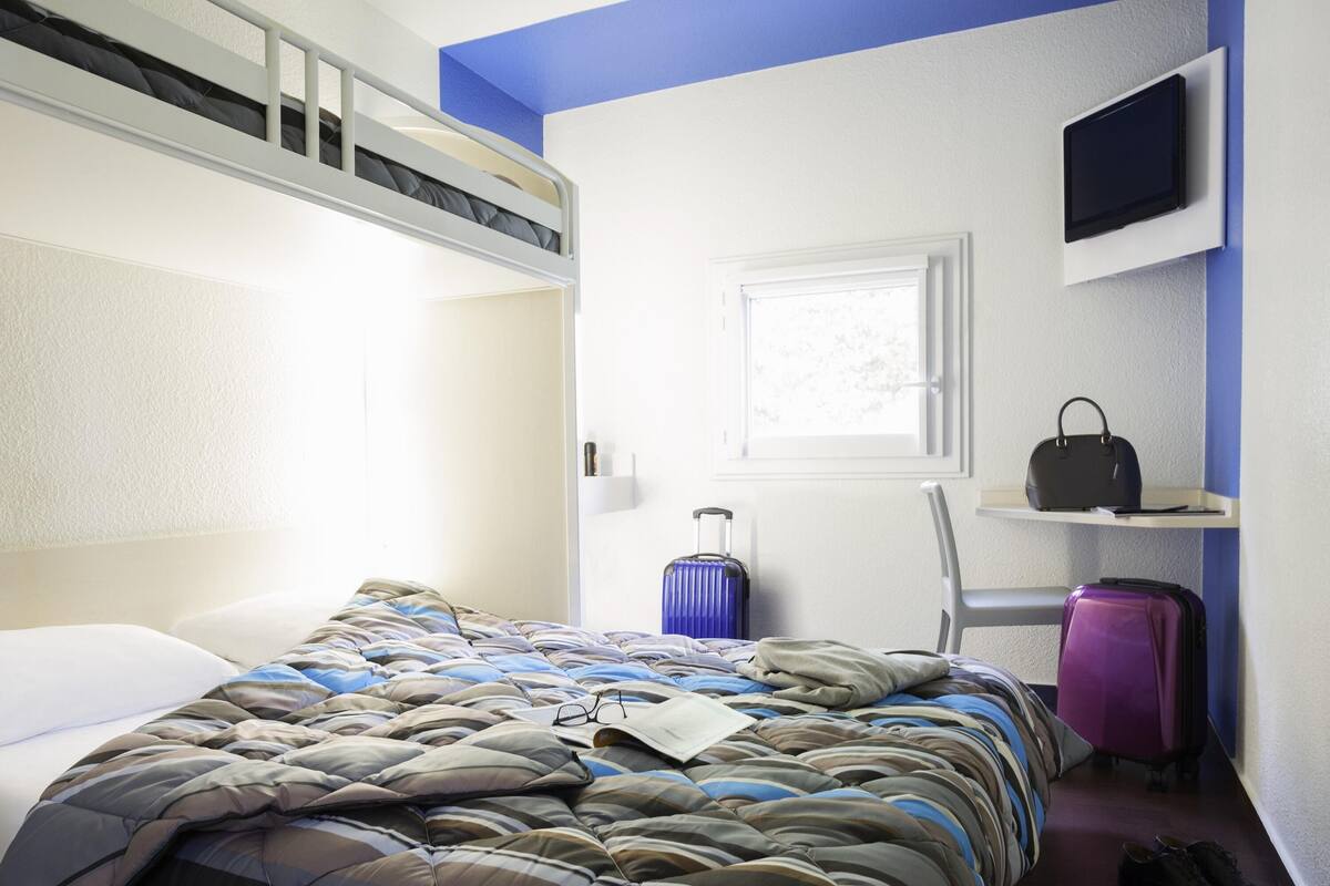 A hotel room with a bunk bed, a suitcase, a chair, a television, and a window.