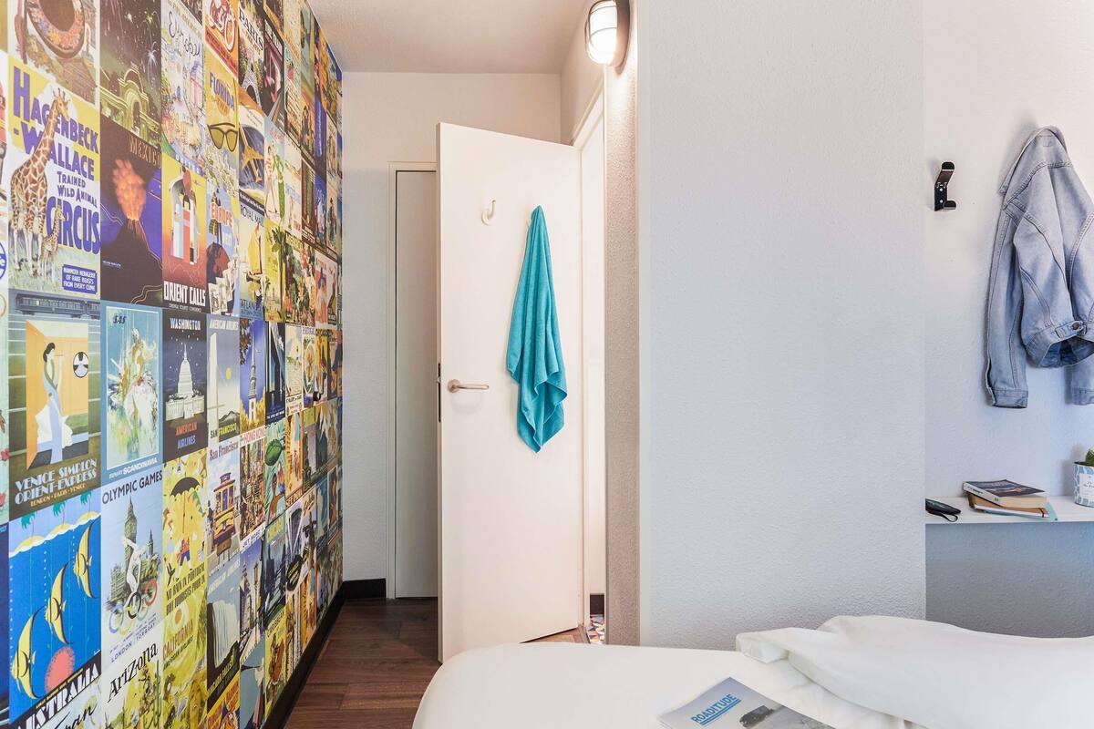 A hotel room with a bed, a door with a towel hanging, a wall with travel posters, and a light fixture.