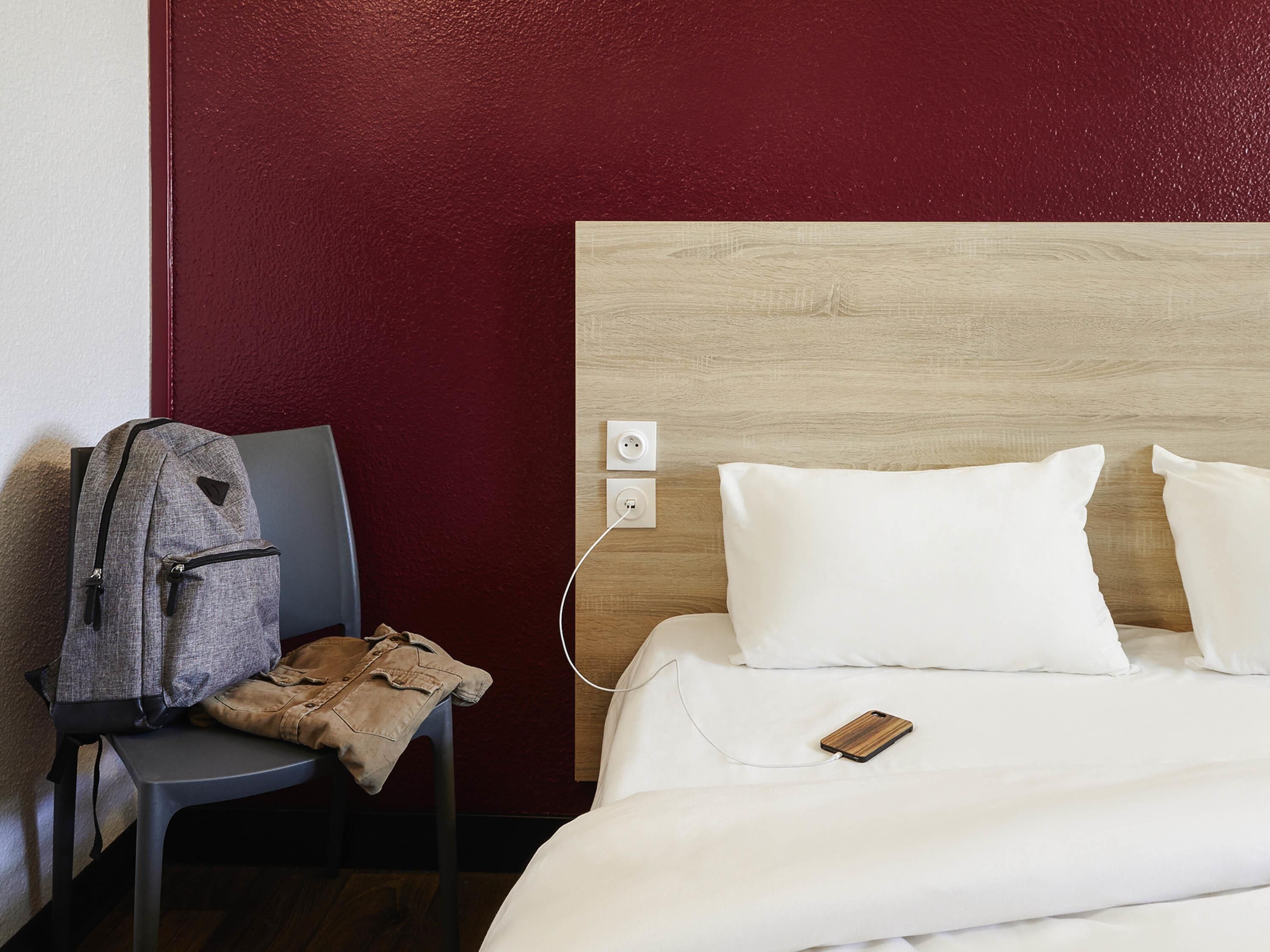 A hotel room with a bed, a chair, a jacket, and a phone charging.