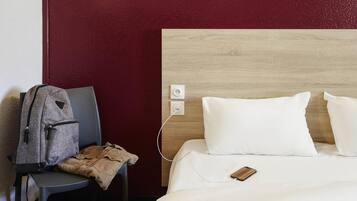 A hotel room with a bed, a chair, a jacket, and a phone charging.