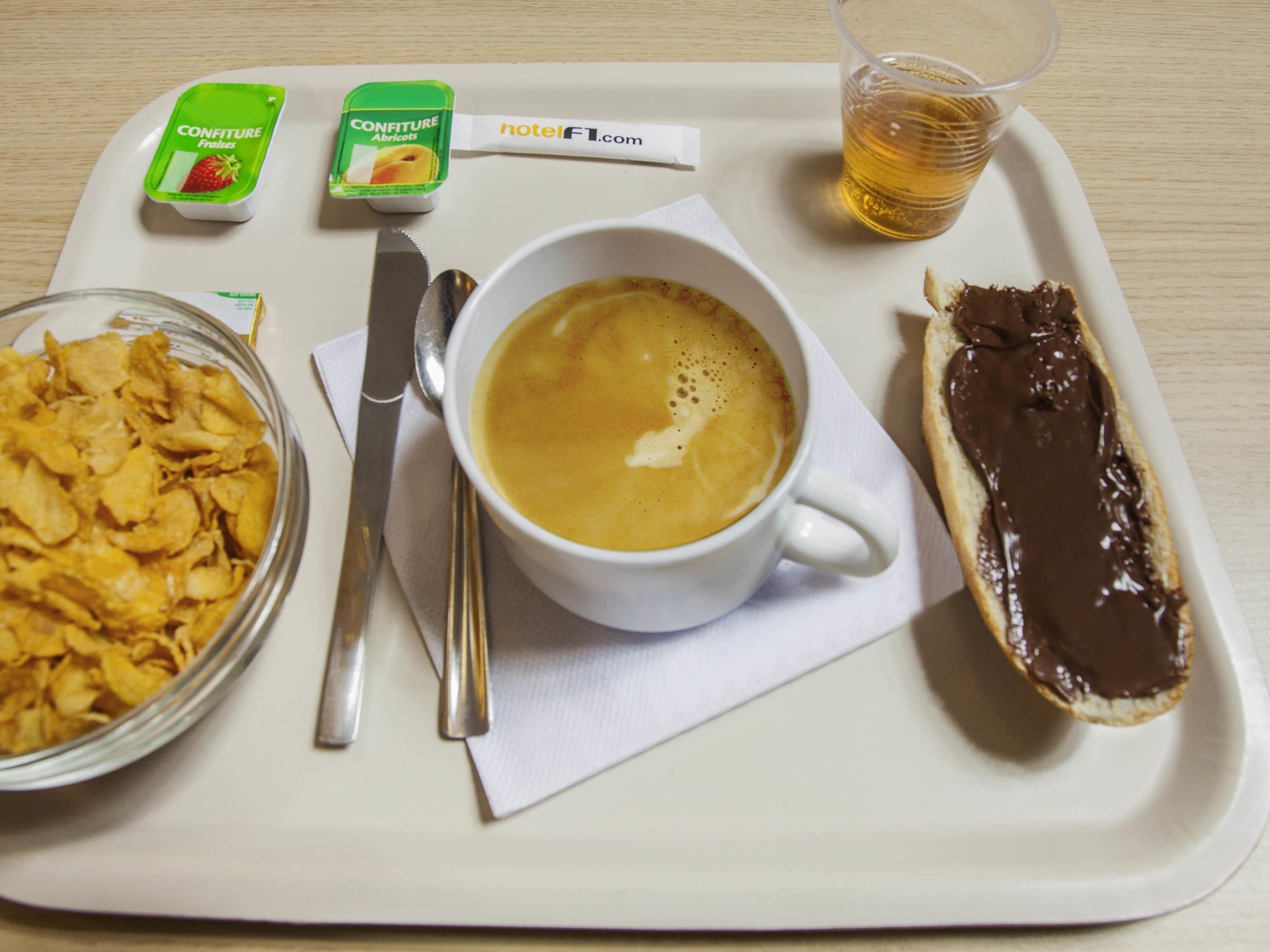 Ein Tablett mit einer Kaffeetasse, einem Schokoladengebäck, einer Schüssel mit Müsli und zwei Joghurtbechern.