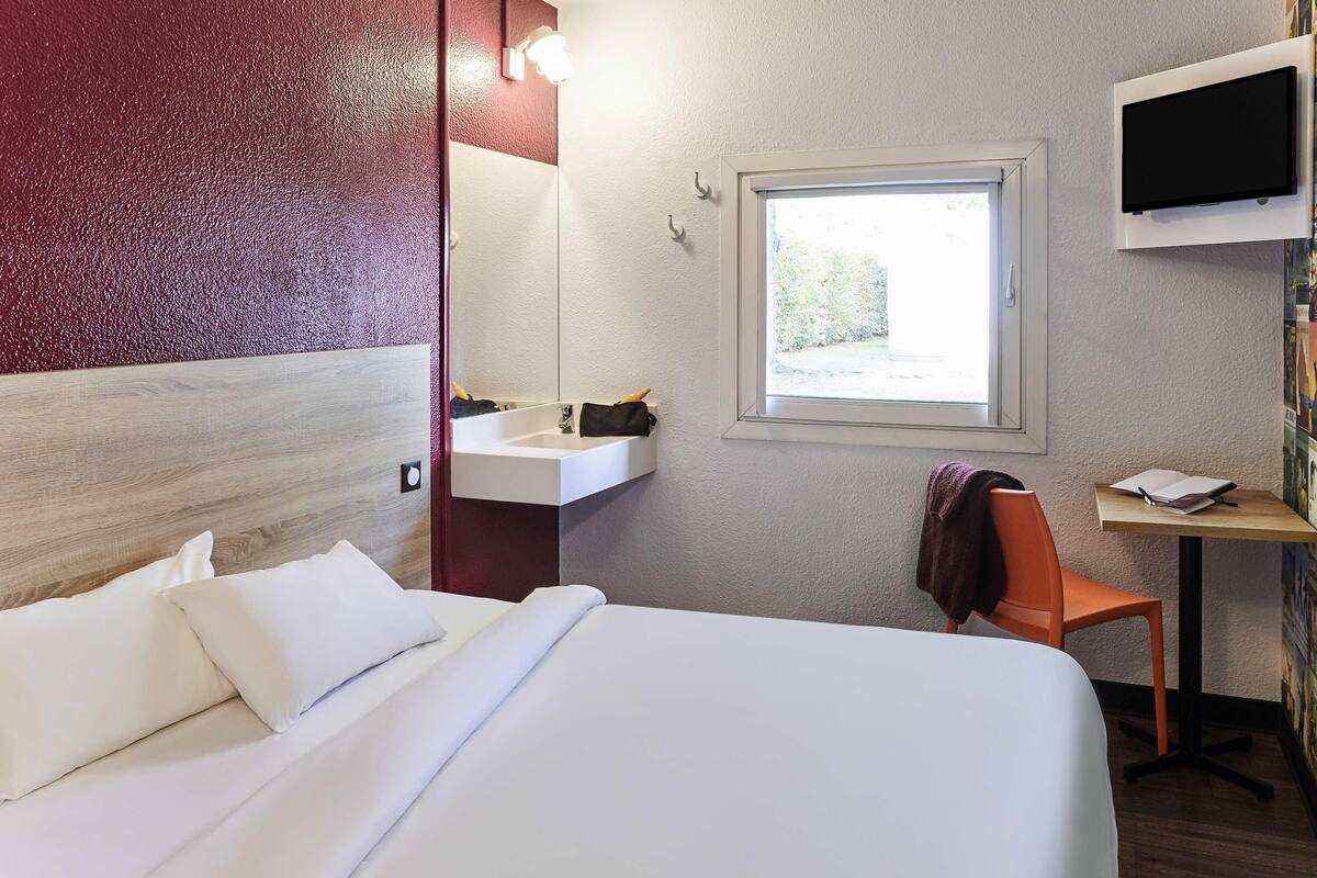 A hotel room with a bed, a desk, a chair, a window, and a TV mounted on the wall.